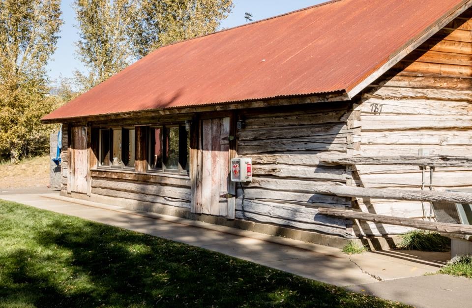 A rustic wooden cabin with a red corrugated metal roof, surrounded by grass and trees. An AED box is mounted on the wall.