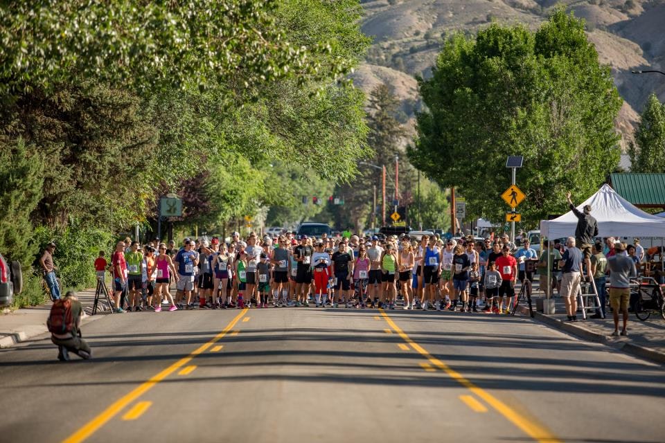 Crowd of runners gathered at the start line of a street race, with lush green trees and hills in the background.
