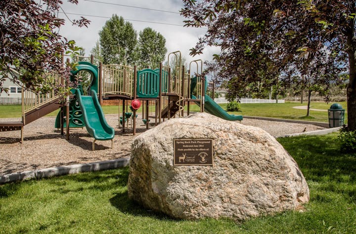 Playground with slides and climbing structures, a large rock with a plaque, trees, grass, and a trash can nearby.
