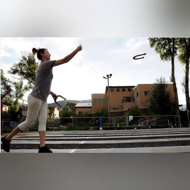 Person tossing a horseshoe outdoors, with buildings and trees in the background.