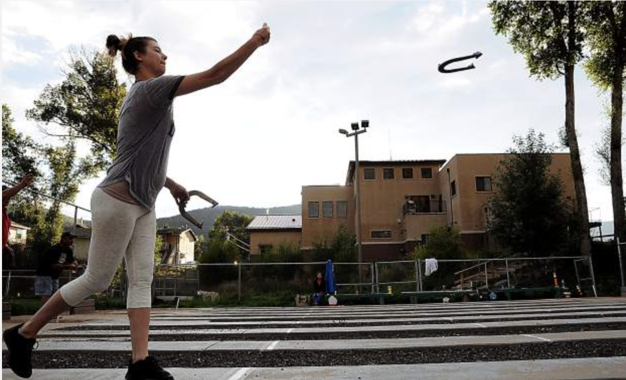 Person tossing a horseshoe outdoors, with buildings and trees in the background.