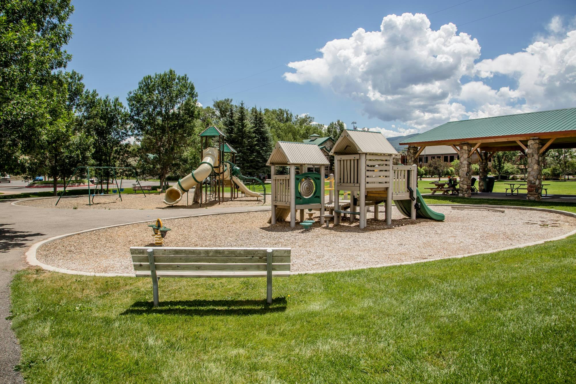 A playground with slides, play structures, swings, a bench, and a pavilion on a sunny day.