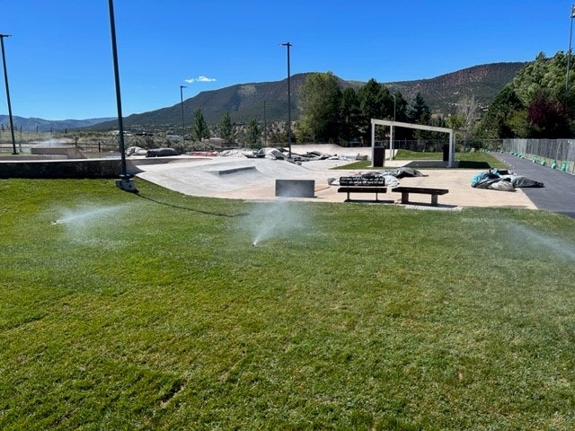 Sprinklers watering grass near a skatepark with ramps and benches, against a background of mountains and trees.