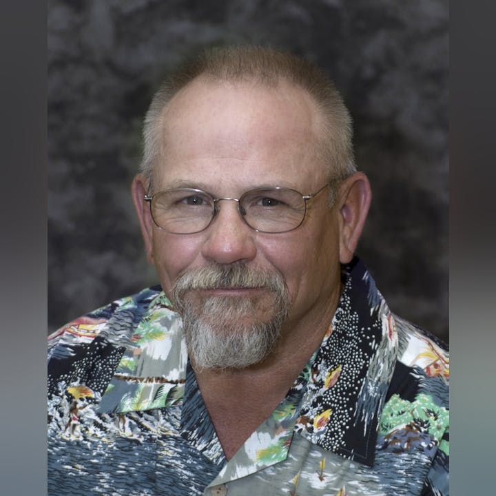 A man with glasses and a beard wearing a colorful Hawaiian shirt poses for a portrait against a dark background.
