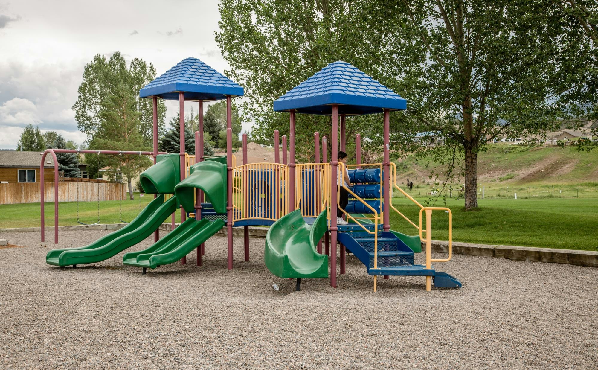 Playground with two slides, climbing structures, and swings, set in a park with trees and grass.