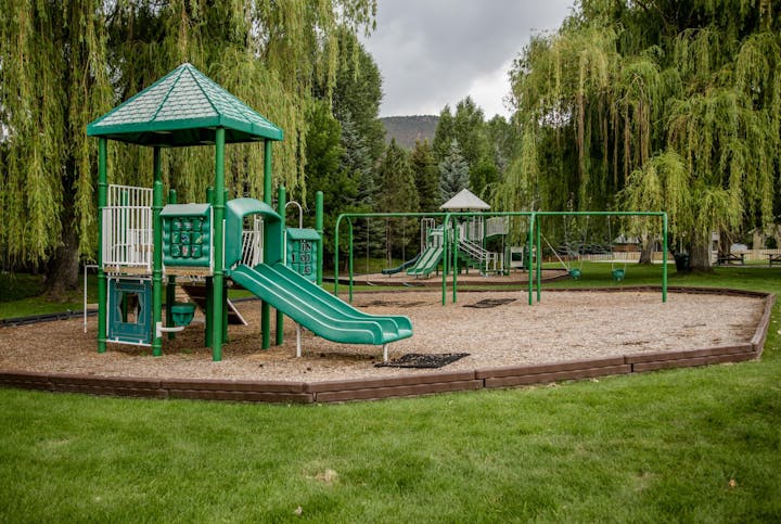 Playground with green slides, swings, and climbing structures. Surrounded by trees and grass.