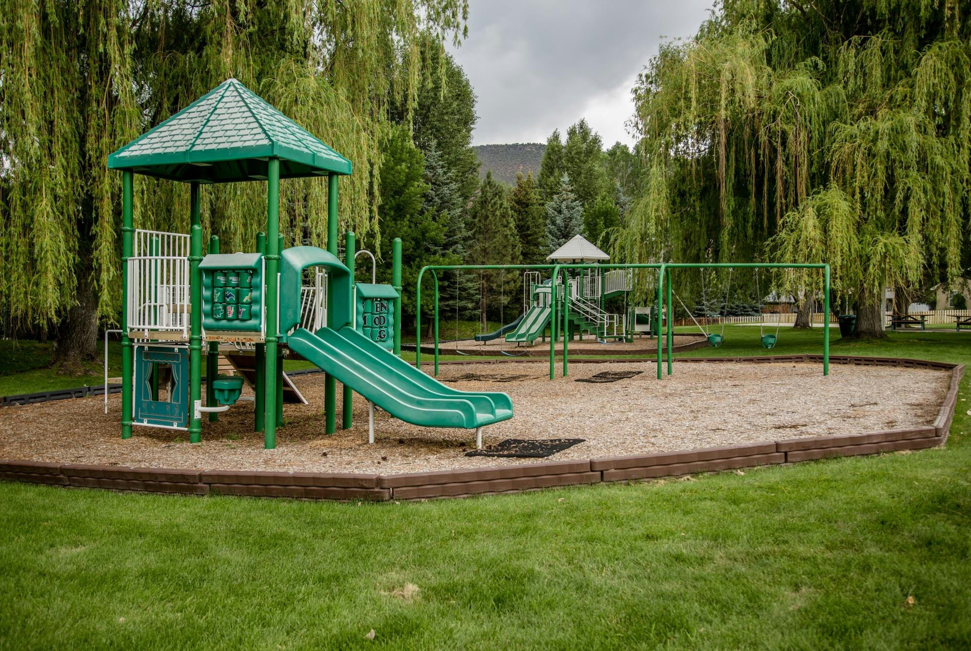 Playground with green slides, swings, and climbing structures. Surrounded by trees and grass.