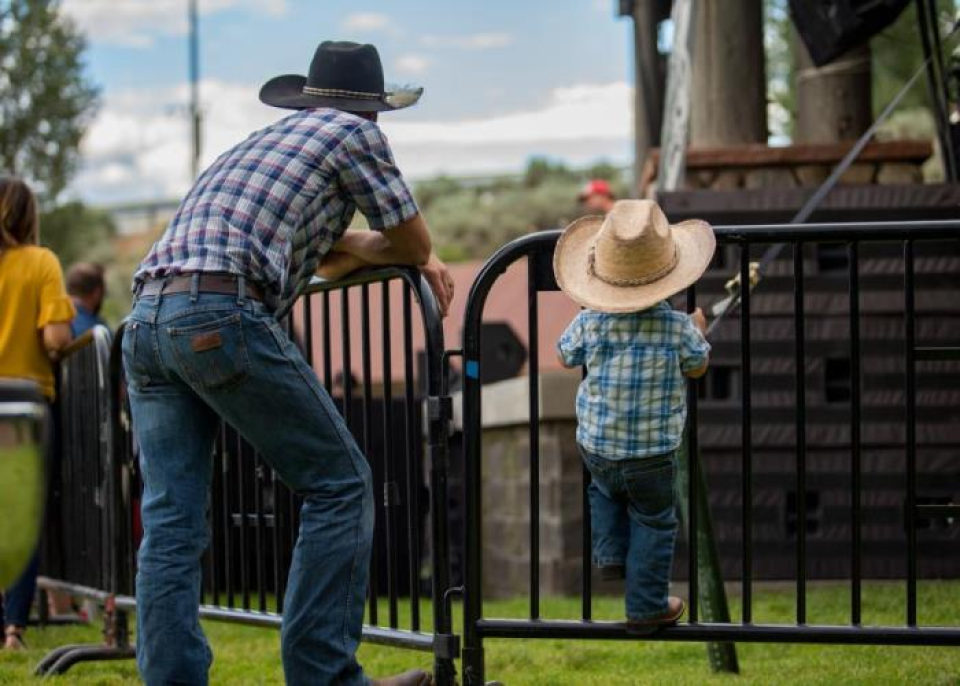 A man and a child in cowboy hats and plaid shirts stand by a fence, watching something outdoors.