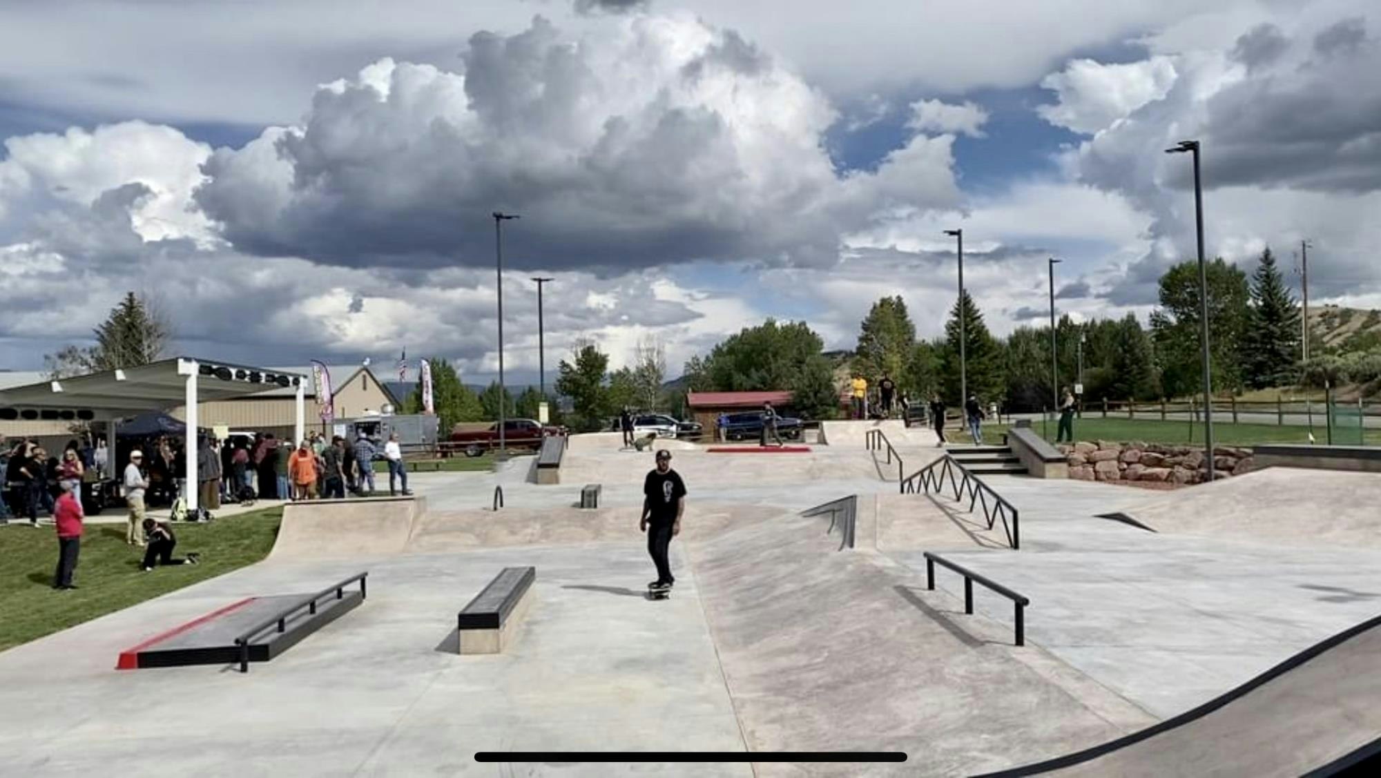 A skate park with people skating and watching, under a cloudy sky.