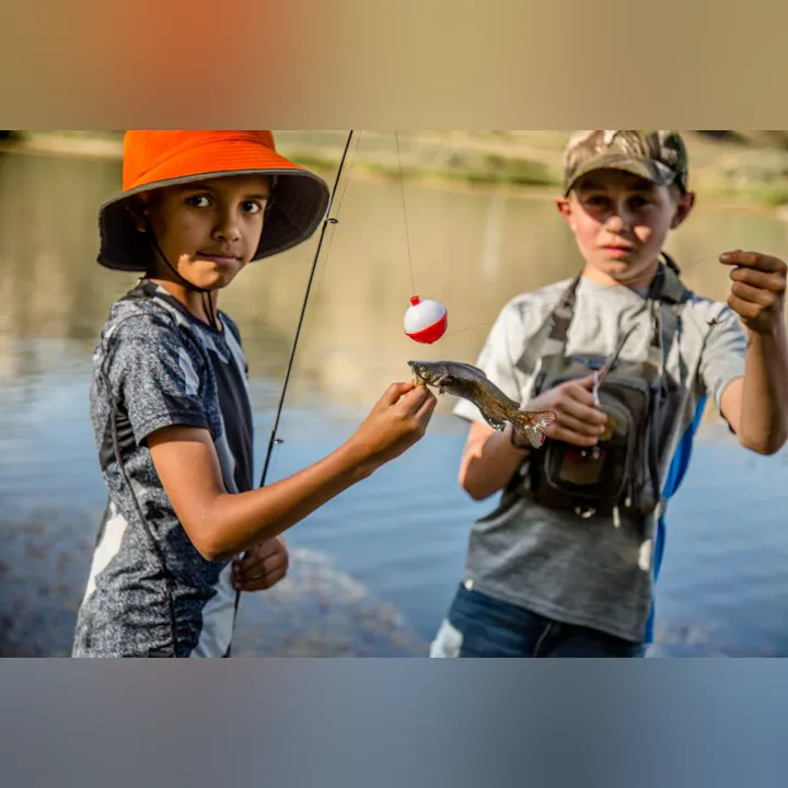 Two kids fishing by a lake. One holds a fishing rod, and the other holds a small fish with a bobber on the line.