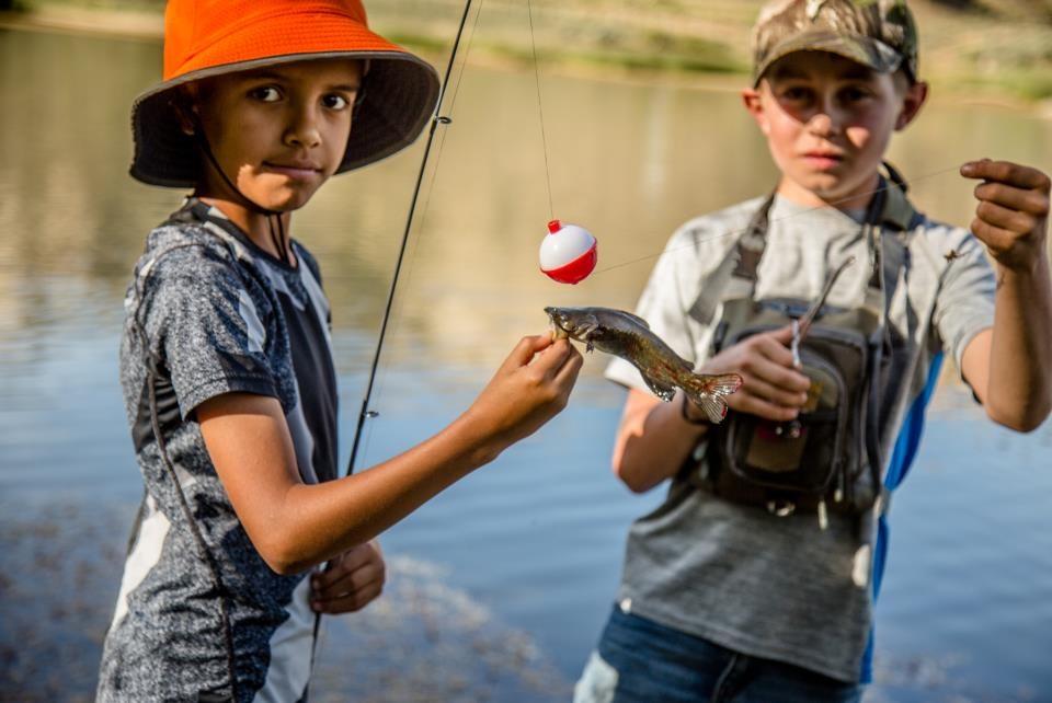 Two kids fishing by a lake. One holds a fishing rod, and the other holds a small fish with a bobber on the line.