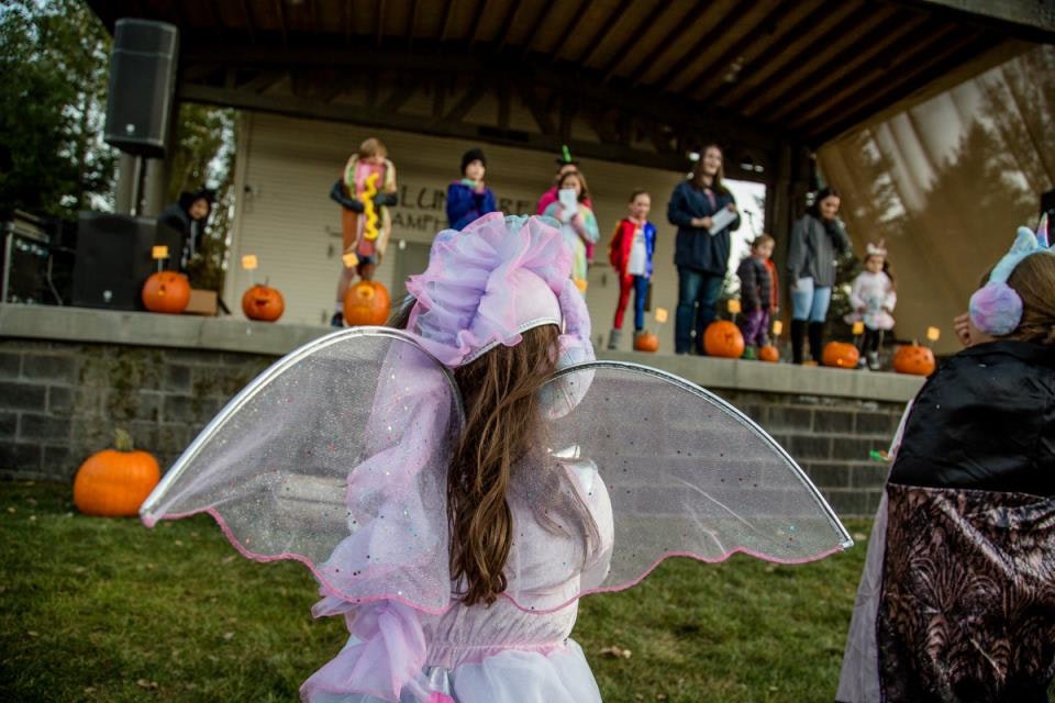A child in a fairy costume with wings looks at a stage with kids and pumpkins.