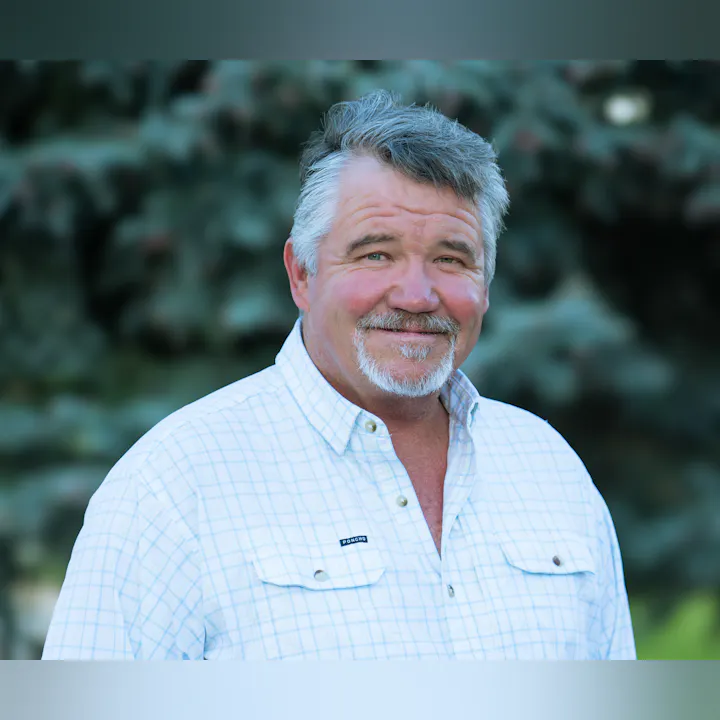 The image shows a smiling man with gray hair, wearing a plaid shirt, standing outdoors with a blurred green background.