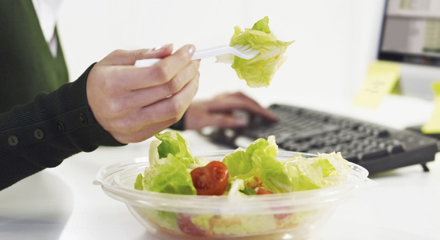 A person eating a salad at a desk with a computer keyboard in the background.