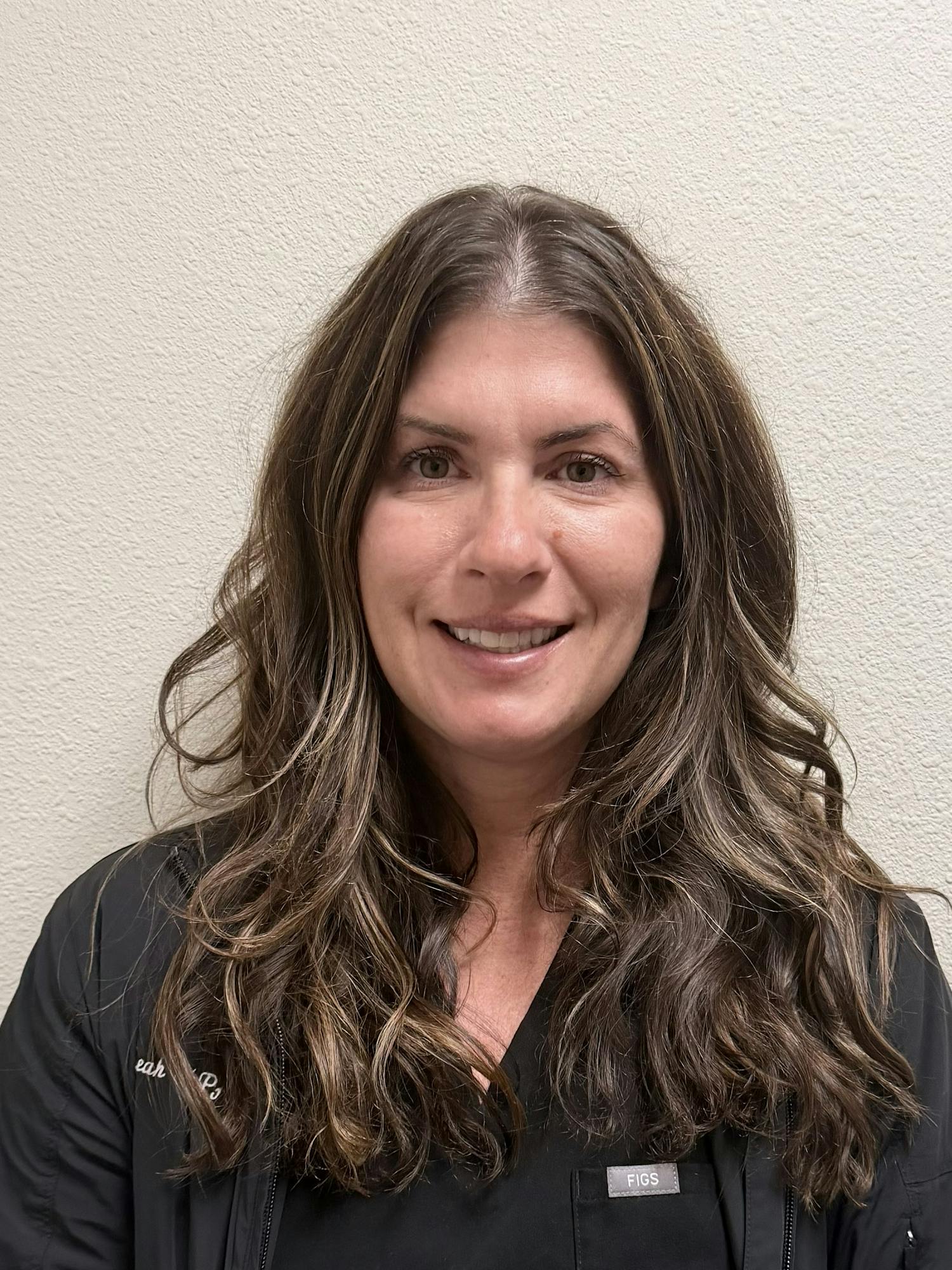 A woman with long hair is smiling, dressed in a black outfit, against a plain light-colored wall.