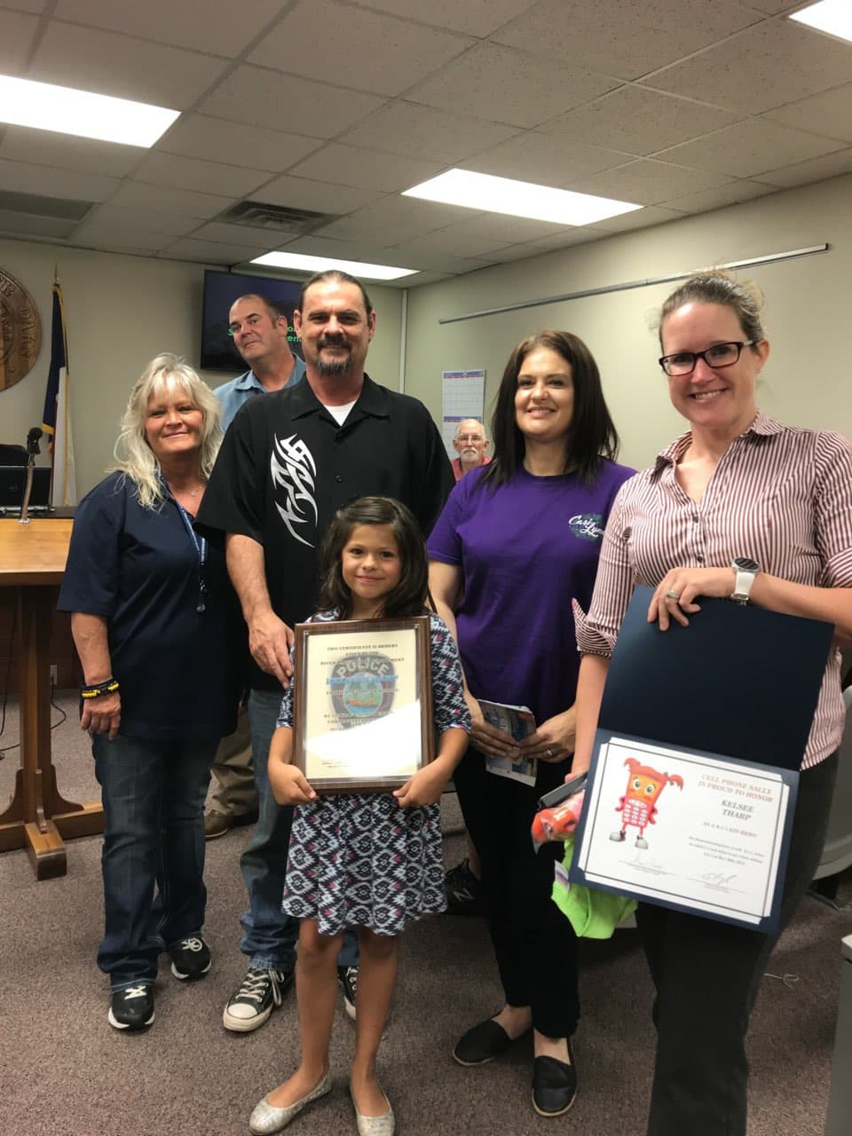 Group of people smiling, including a young girl holding a certificate, in an indoor setting possibly at an award ceremony.