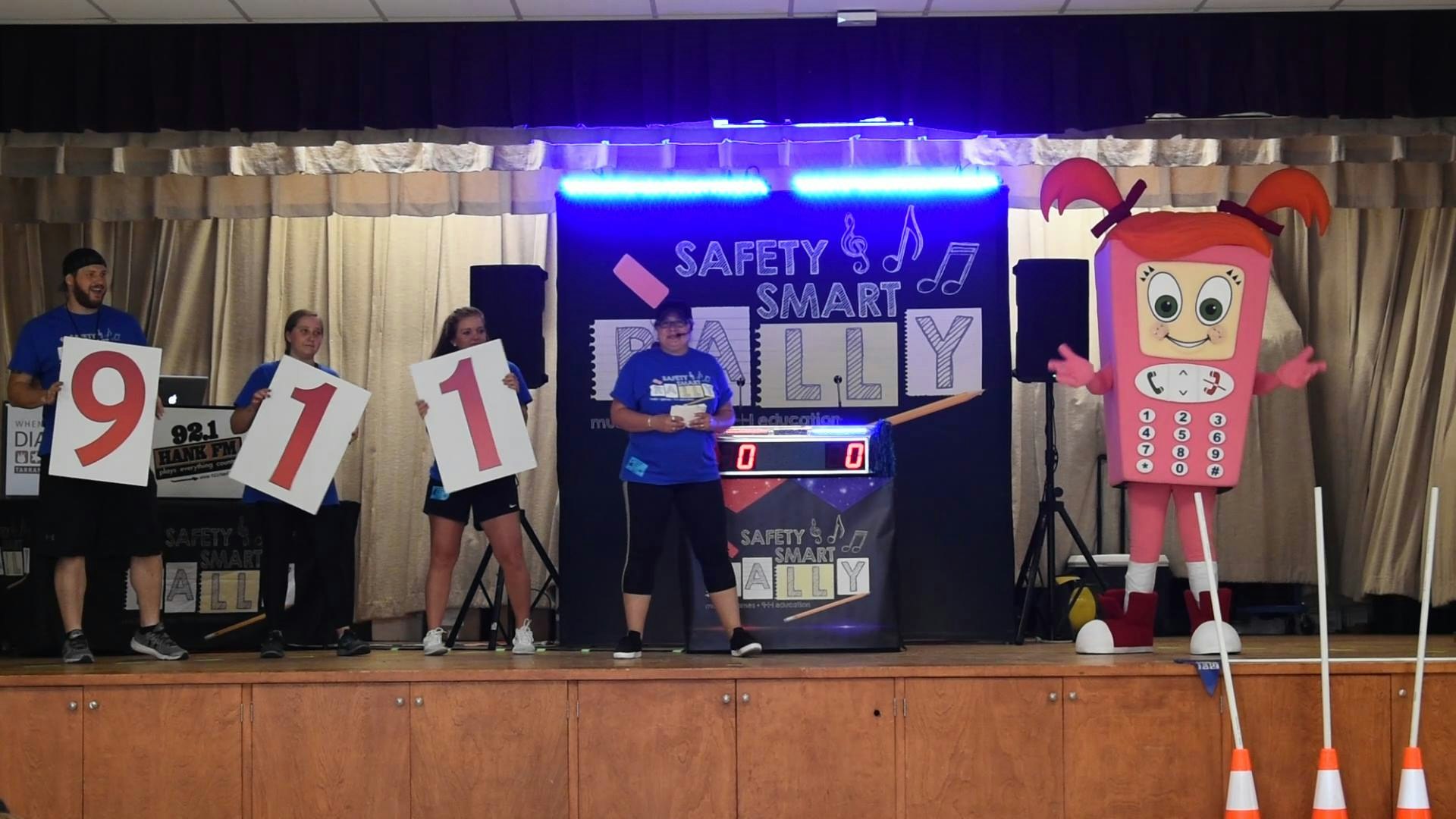 People with "9-1-1" signs, a mascot resembling a phone, and a "Safety Smart Billy" backdrop at an event.