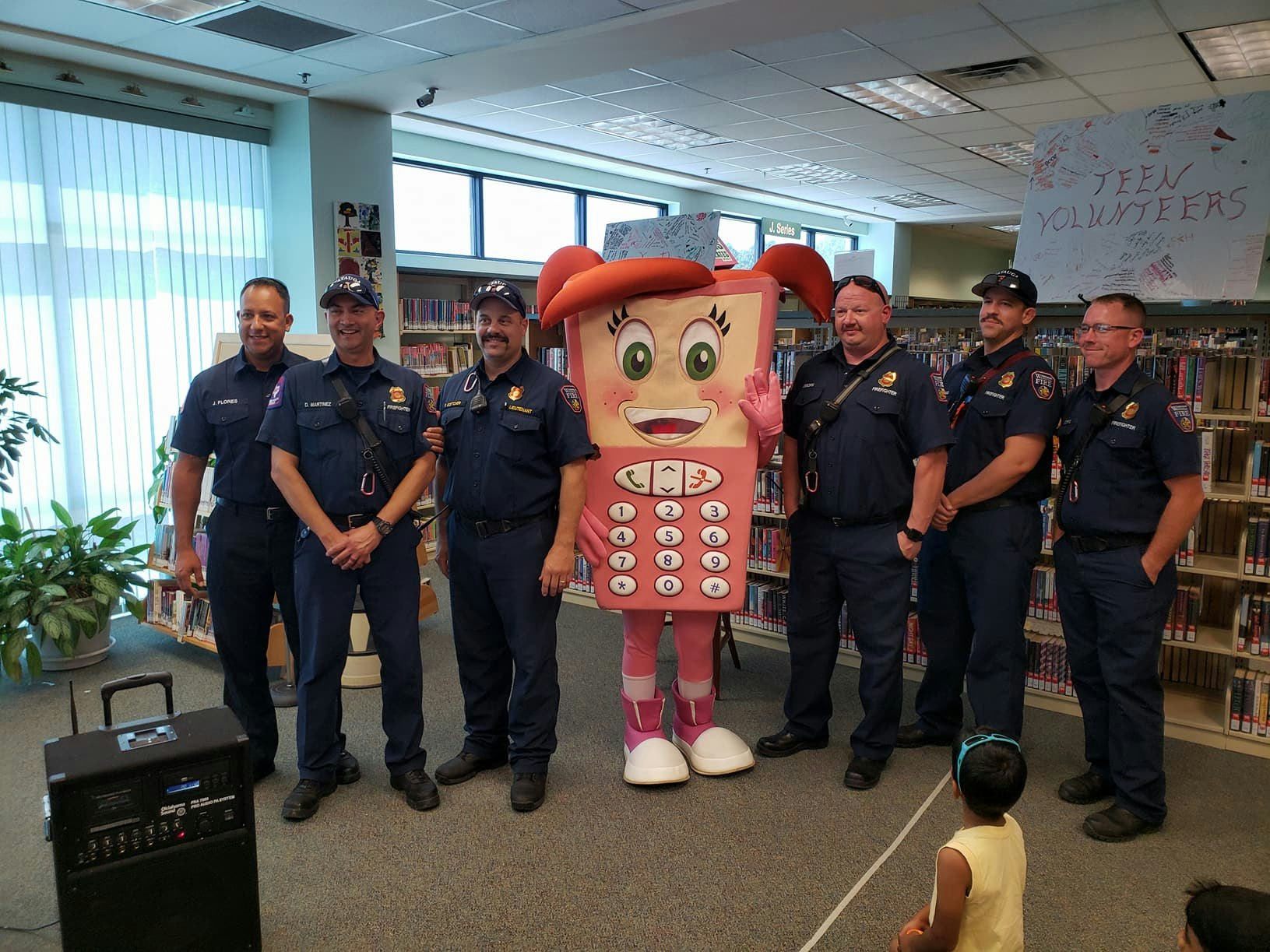 Firefighters stand with a mascot resembling a phone, with a child looking on at an indoor event.