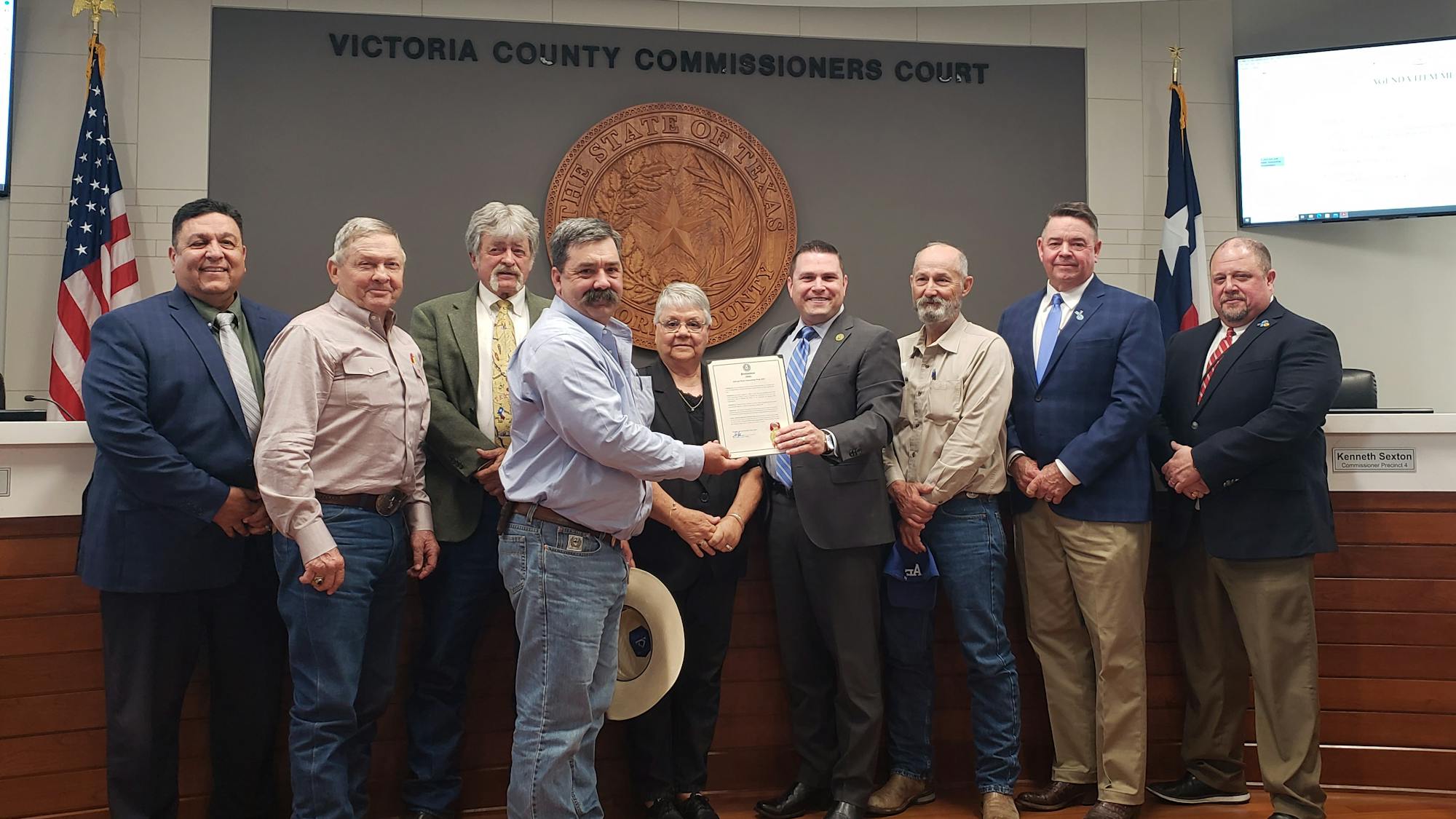 A group of people standing in front of the Victoria County Commissioners Court seal, with two individuals holding a certificate.