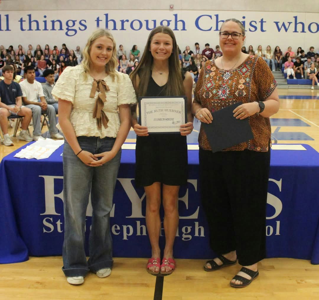 Three people standing in a gym; middle person holding a certificate. Audience seated in background.