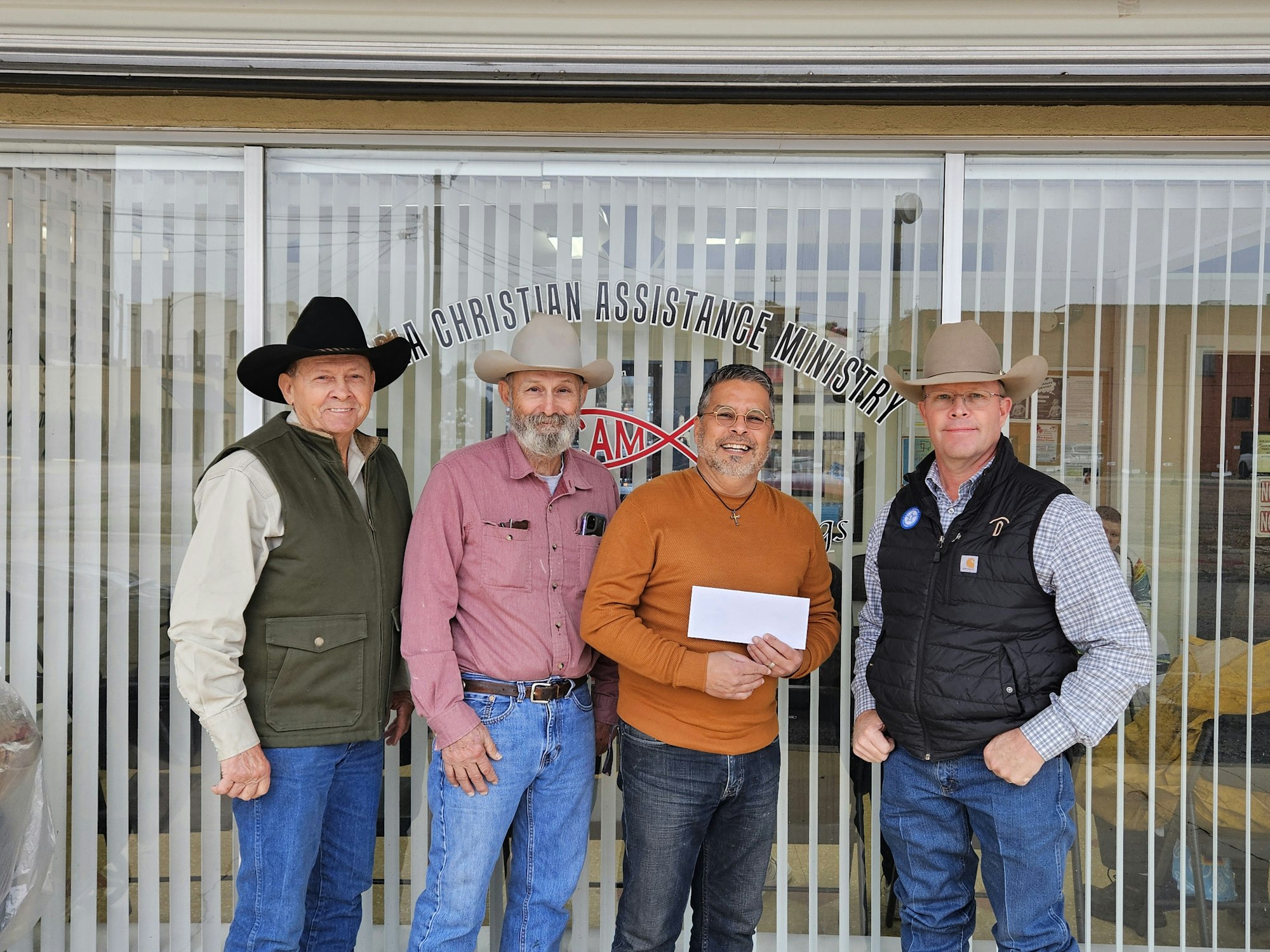 Four men stand in front of a building with "Christian Assistance Ministry" on the window, smiling and posing for a photo.