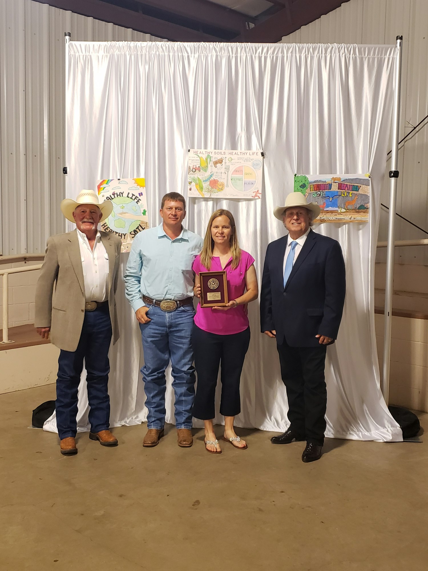 Four people standing together, one holding a plaque, in front of agriculture-themed posters with a white curtain backdrop.