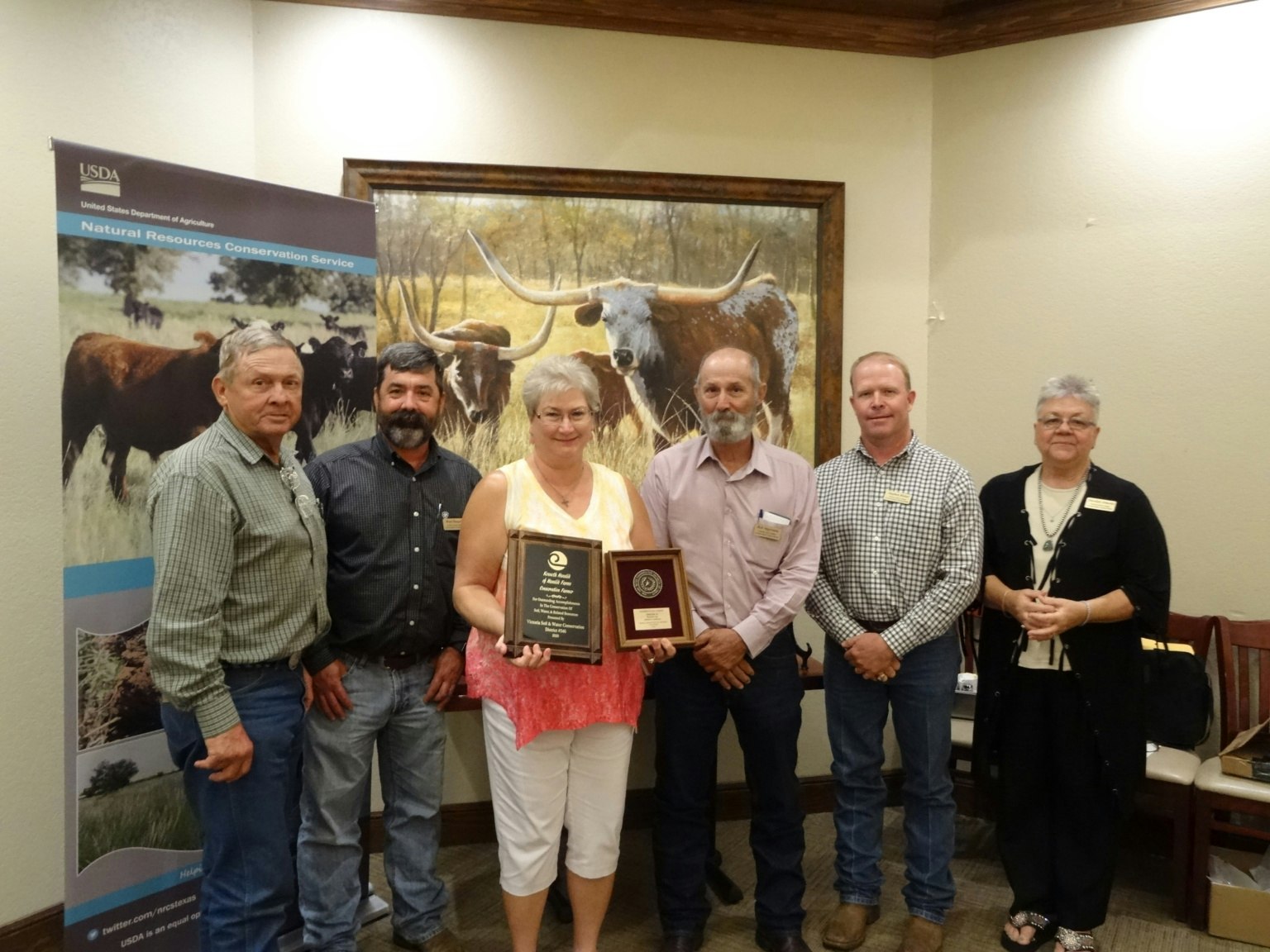 A group of six people poses with plaques in front of a USDA banner and a cattle painting.