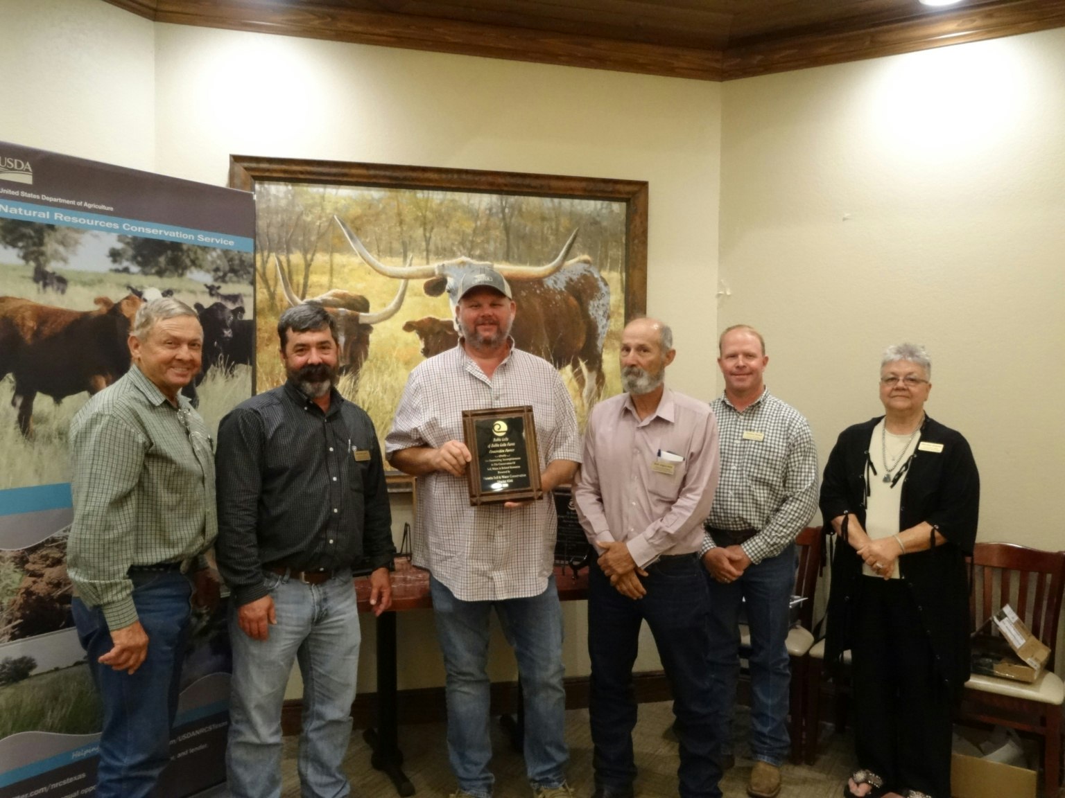 A group of six people stand together; one man holds a plaque. A banner and nature painting are in the background.