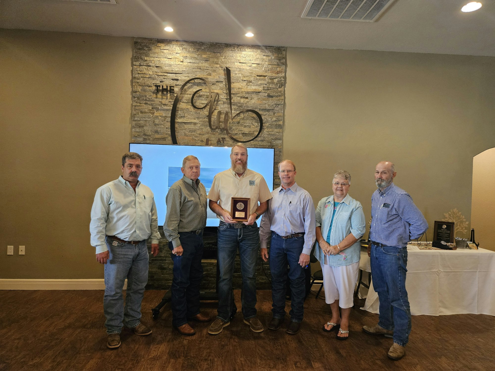A group of people stands together indoors, with one person holding a plaque. They're in front of a sign that says "The Club."