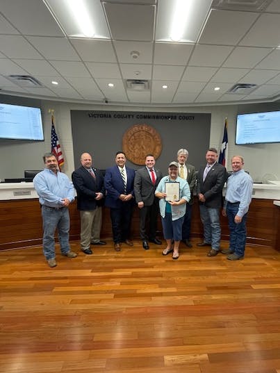 A group of people in a meeting room, one holding a certificate. Flags and monitors are in the background.