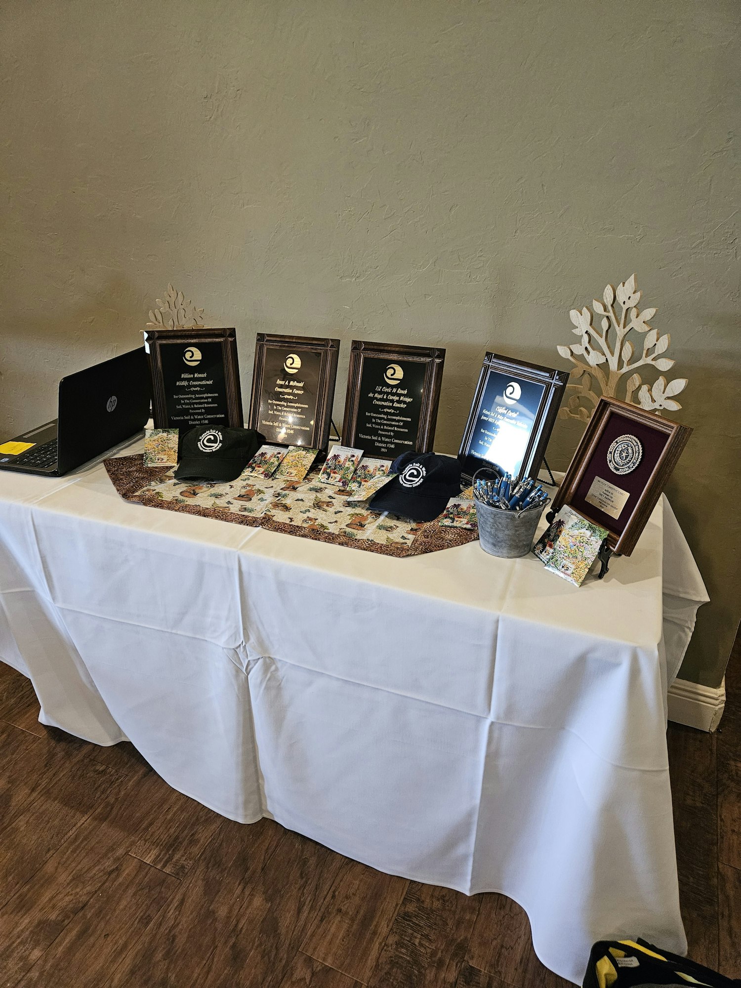 A table with awards, a laptop, hats, and brochures on a tablecloth.