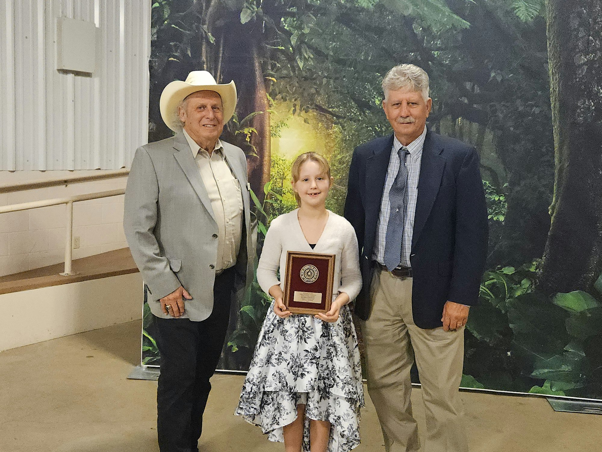 Three people posing indoors; a young girl holding a plaque, flanked by two men in suits. Background features a forest scene.