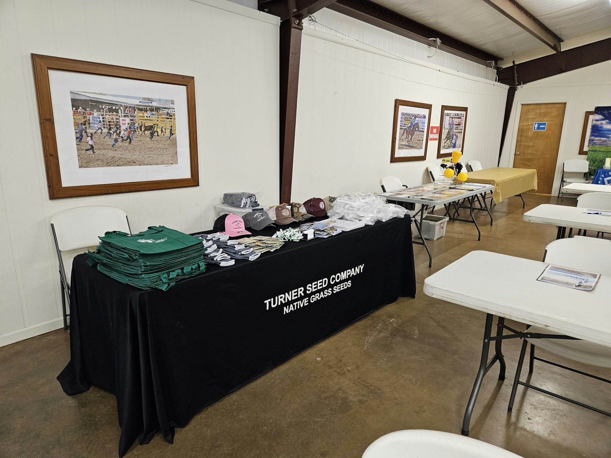 Promotional table with Turner Seed Company items like bags, hats, and shirts in a room with framed photos and tables.