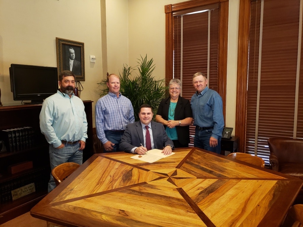 Five people in an office, one signing a document at a wooden table with a star design.