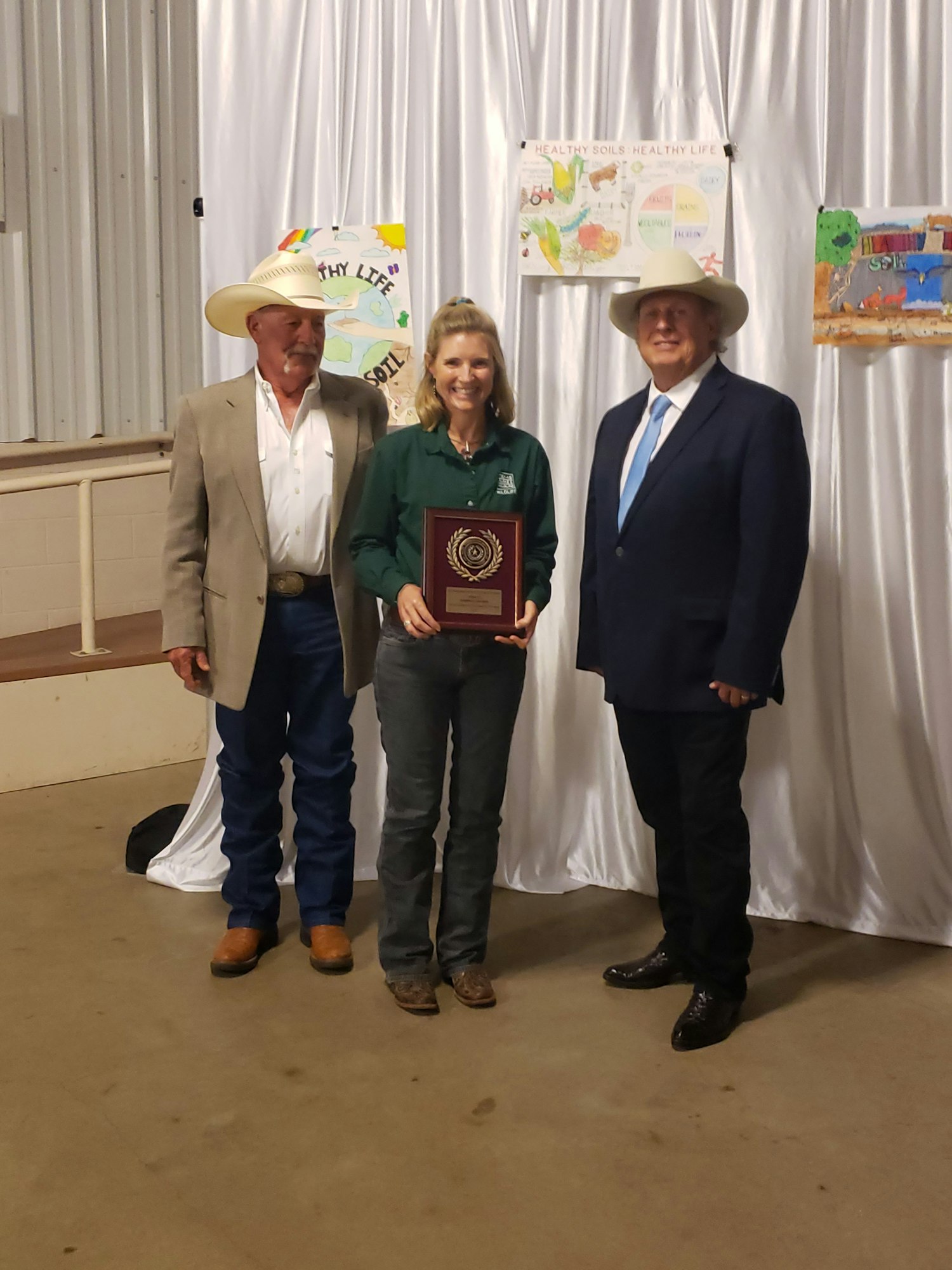 Three people posing, one holding an award plaque, with posters about healthy soil behind them.