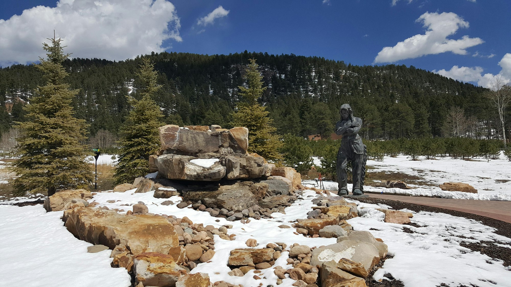 Statue by a rocky stream, evergreen trees, and a snowy landscape with forested hills in the background.