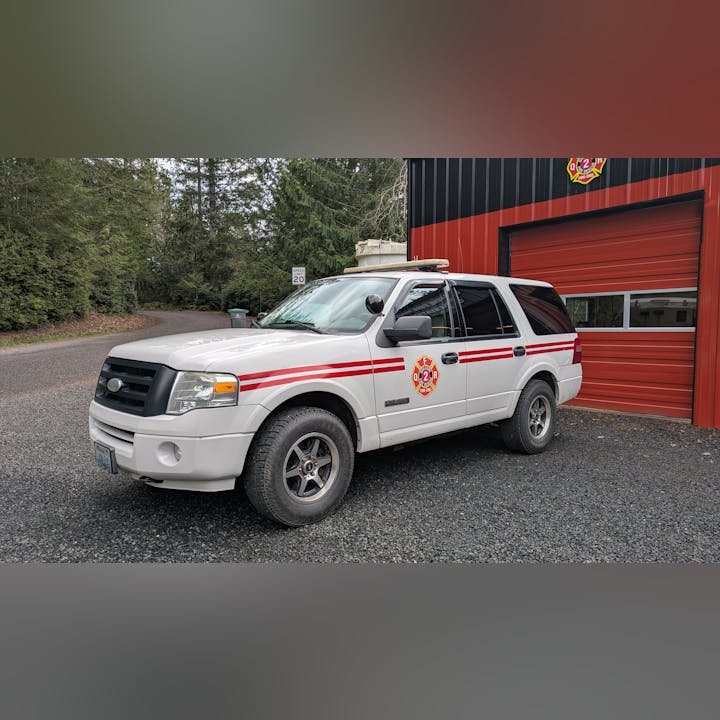 A white fire department SUV with red stripes is parked beside a red building, near a speed limit sign, in a forested area.