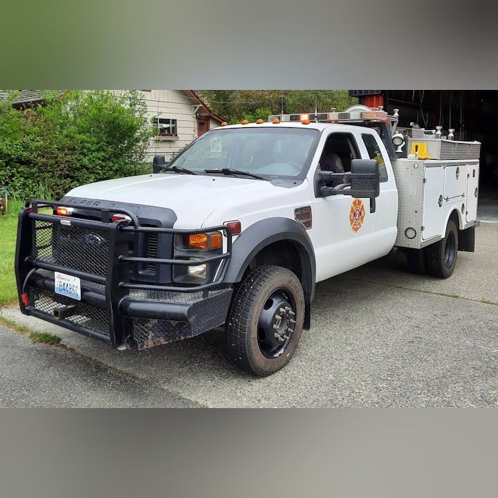 The image shows a white Ford truck with a fire department logo, equipped with emergency lights and firefighting gear in the back.