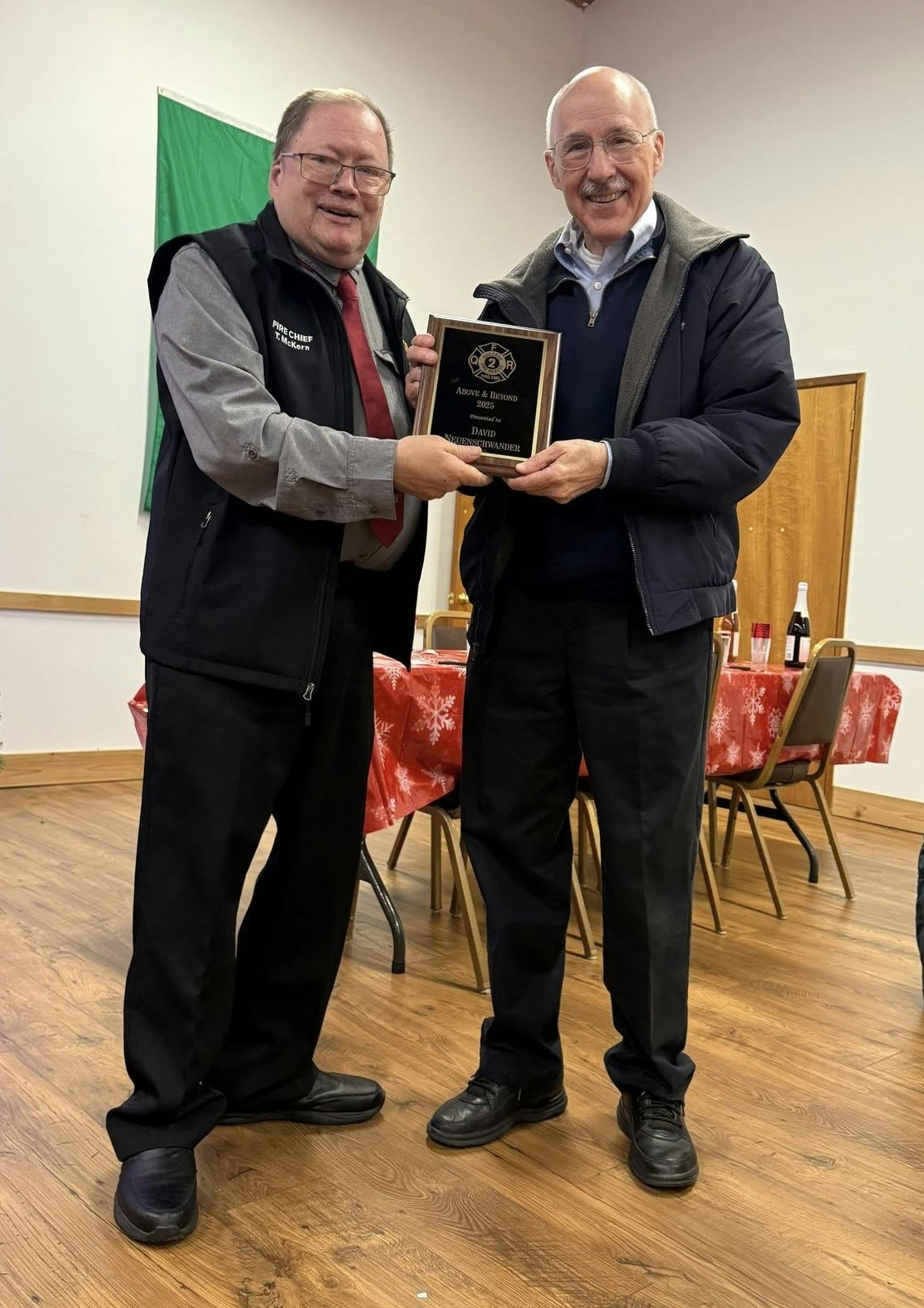 Two men are presenting an award plaque to one of them at a festive event, with holiday-themed decorations in the background.
