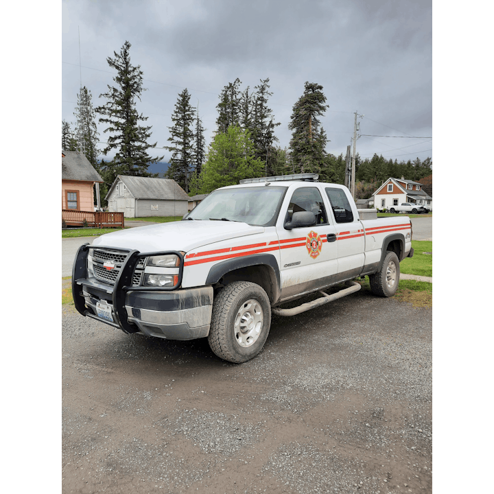 The image shows a white Chevrolet pickup truck with fire department markings, parked alongside a gravel area near trees.