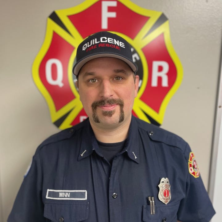 A firefighter in uniform stands in front of a Quilcene Fire Rescue emblem.