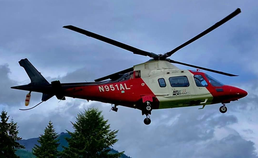 A red and white medical helicopter mid-flight against a cloudy sky with trees below.