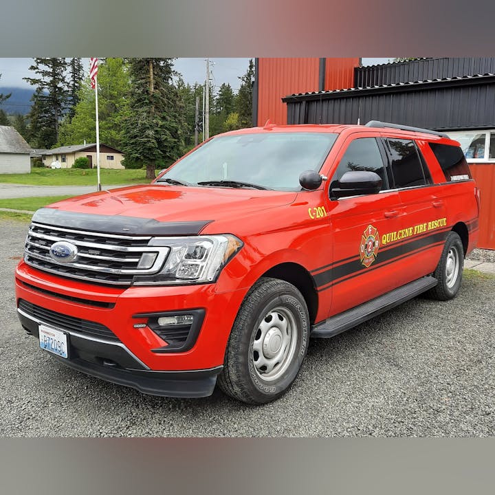 A red Ford SUV marked as "Quilcene Fire Rescue," parked in a rural area with trees and a building in the background.