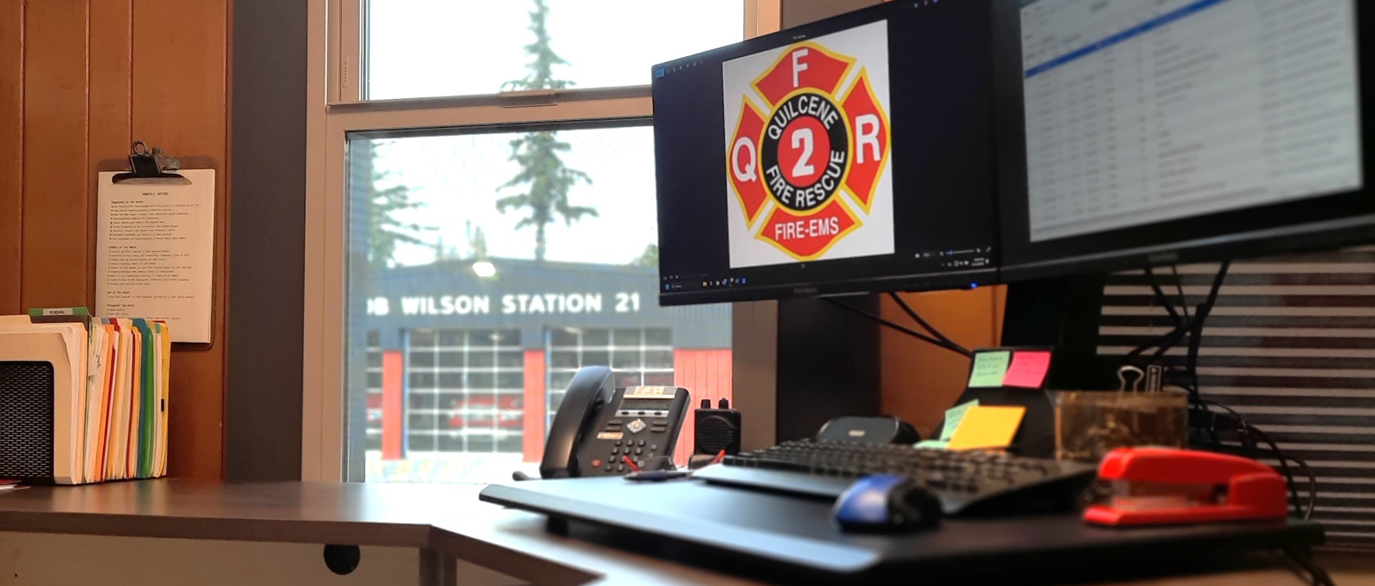 The image shows a workspace with a computer displaying a fire department logo, folders, a phone, and a view of a fire station outside.