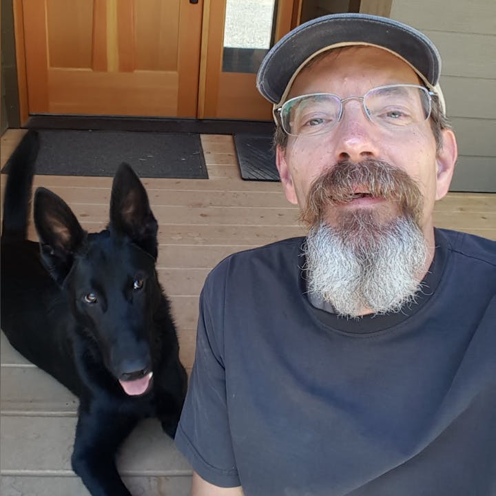 A man with a beard and glasses takes a selfie with a black dog on a wooden porch.