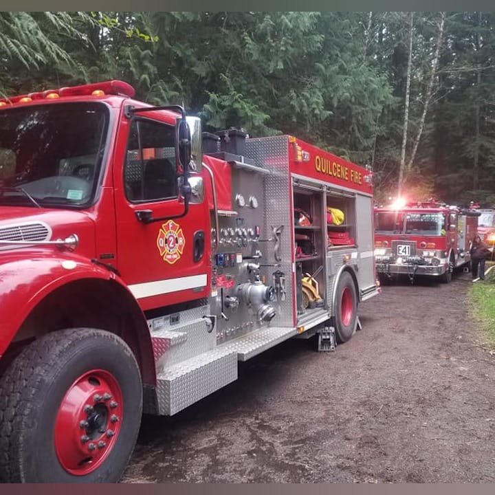 The image features two fire trucks, one prominently labeled "Quilcene Fire," parked in a wooded area.