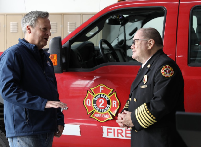 Two men are talking near a fire truck, one in a jacket and the other in a fire chief uniform.
