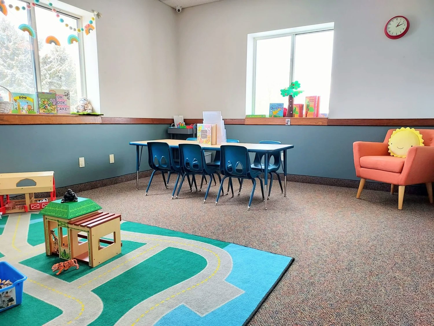 A colorful children's playroom with a table, chairs, books, toys, and a cozy chair with a sun pillow.