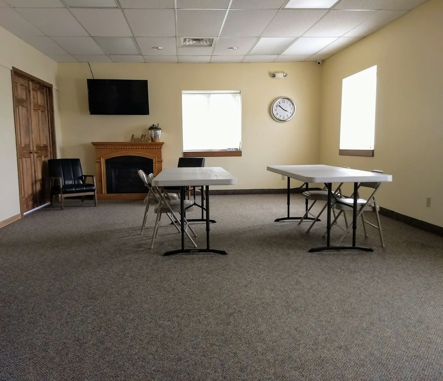 A small, empty room with foldable tables, chairs, a wall clock, TV, fireplace, and windows letting in natural light.