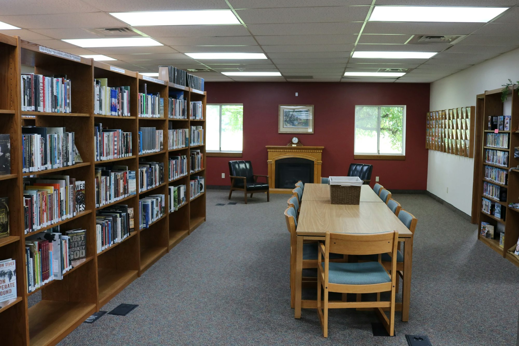 Library interior with bookshelves, tables, chairs, a fireplace, windows, and a maroon accent wall.